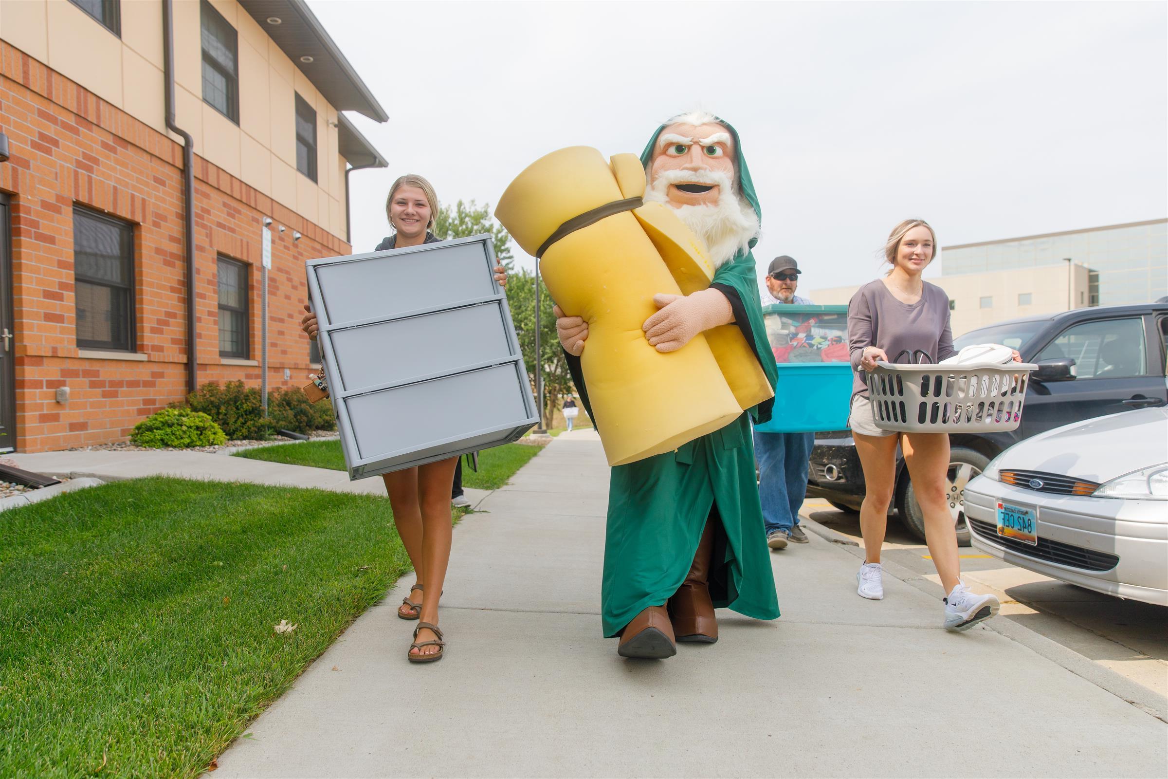 Merlin the Mystic Mascot is helping 2 smiling female students move in to the residence hall